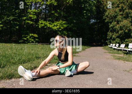 Young Woman Stretching Legs on Asphalt Before Running in Sunny Park. Stock Photo