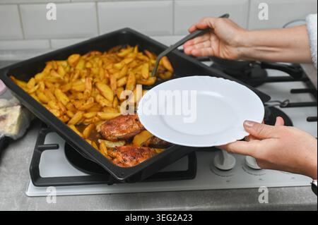 Woman holds empty white plate over baking tray with roasted potatoes. Woman prepares to serve homemade dinner from baking tray. Hot chicken and potato Stock Photo