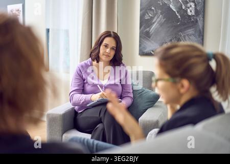 Female therapist attentively listening and taking notes as couple discusses relationship concerns during therapy meeting, calm counseling room interio Stock Photo