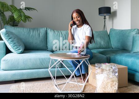 Smiling woman talks on her phone and writes notes at a coffee table while packing resale orders with cardboard boxes at home, organizing shipments for Stock Photo