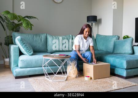 Black woman in a modern living room packing a secondhand marketplace order into a cardboard box, preparing shipment for an online app sale, focused on Stock Photo