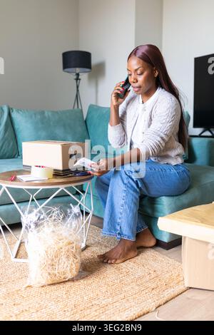 Young woman sitting on a sofa making a phone call while checking a parcel and paperwork, organizing home delivery items in a modern living room. Stock Photo