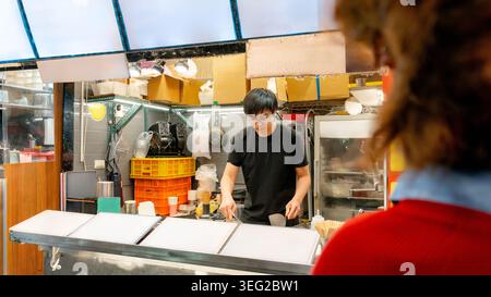 Taiwanese street food vendor cooks at a bustling night market stall, with bright lights, utensils, and takeaway cups, capturing local cuisine culture Stock Photo