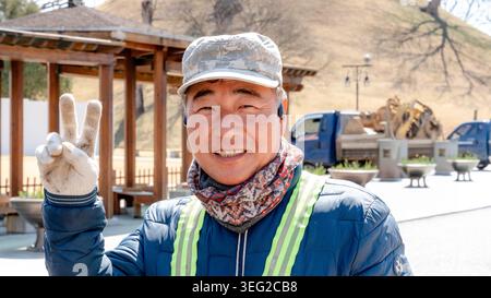 A road repair worker in Gyeongju, South Korea, smiling and gesturing during a break. The backdrop features construction equipment and a serene hillsid Stock Photo