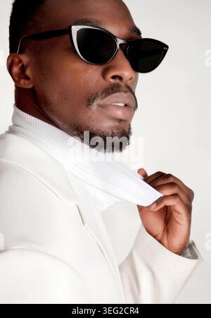 Closeup portrait of a handsome Black man in an elegant white outfit and dark sunglasses, adjusting his collar in a studio setting with clean backgroun Stock Photo