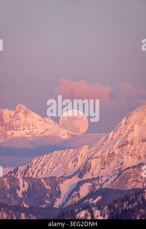 The full moon rises majestically behind the snow-capped peaks of Dents du Midi in the Alps during a serene sunset, bathing the landscape in a warm gol Stock Photo