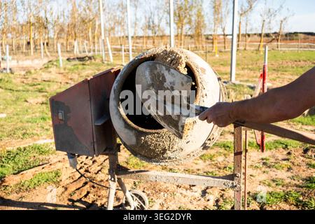 Close-up of a construction worker using a shovel to load wet concrete into a rotating cement mixer outdoors, showing building work equipment and dusty Stock Photo