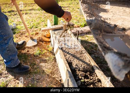 Worker pours wet concrete into wooden formwork for an exterior foundation or curb, using tools to spread and level the mix during a construction site Stock Photo
