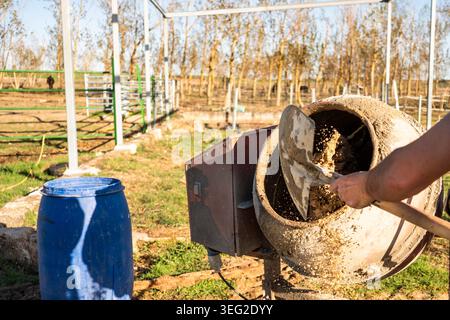 Close-up of worker using a shovel to load wet concrete into a rotating mixer at an outdoor construction site, with metal frame structures and a blue b Stock Photo