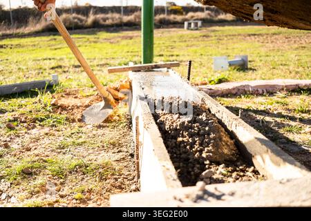 Worker pours and levels wet concrete in wooden formwork outdoors, shaping a small foundation or curb. Exterior construction site with shovel, fresh ce Stock Photo