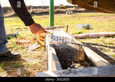 Close-up of a worker leveling wet concrete in a wooden form outdoors, using a trowel for foundation edging during construction work on an exterior sit Stock Photo