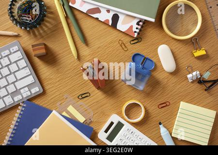 Contemporary home office flat lay with keyboard, notebooks, pens, clips, tape, calculator and sticky notes on wooden desk, ideal for productivity, stu Stock Photo