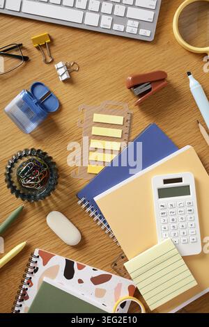 Contemporary workspace flat lay on wooden desk featuring keyboard, notebooks, calculator, sticky notes, clips and pen, creating a productivity-focused Stock Photo