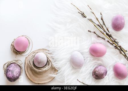 Minimal Easter composition with pastel purple and pink eggs arranged on soft white fur, nestled in natural nests and paired with a budding spring bran Stock Photo
