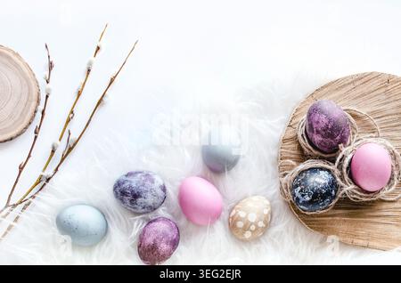 Pastel Easter egg composition on soft white fur with rustic wooden board, willow branches, and speckled dyed eggs in purple, pink, blue, and beige. Stock Photo