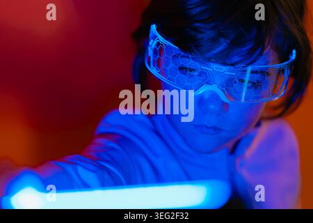 Close-up of a kid girl wearing smart glasses, reaching toward a glowing light bar in neon blue and red studio lighting, showcasing futuristic technolo Stock Photo