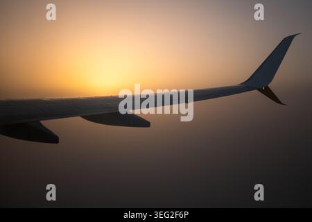 Airplane wing in silhouette during golden sunrise, viewed from a passenger window above a soft cloud layer. Calm travel mood, aviation concept, and se Stock Photo