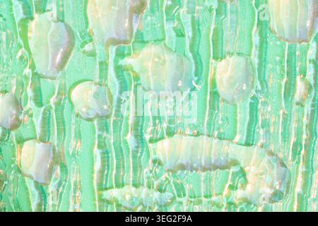 Close-up of cosmetic serum drops and streaks on textured glass, illuminated by neon green and blue lighting. Abstract skincare background with glossy Stock Photo