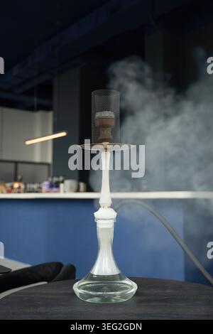 A decorative hookah stands on a dark table in a modern bar, with ambient lighting and soft clouds of smoke, evoking a sense of relaxation and style. Stock Photo