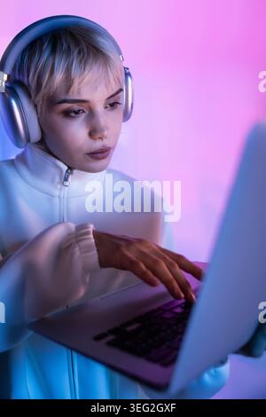 Futuristic woman in white jacket wearing headphones, focused on a laptop in pink and blue neon light. Modern cyber aesthetic, technology concept, nigh Stock Photo