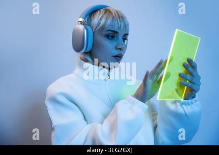 Futuristic woman in a white jacket wears wireless headphones while interacting with a glowing green tablet, bathed in blue light, exploring digital in Stock Photo