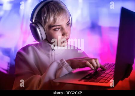 Futuristic woman wearing headphones and a white jacket works on a laptop, focused on typing in a neon pink and blue glow, creating a modern cyberpunk Stock Photo