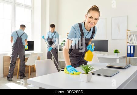 Smiling cleaner wiping office desk with spray bottle as team sanitizes workspace Stock Photo