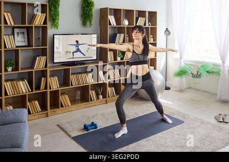Woman doing online yoga workout at home, warrior pose training in living room Stock Photo