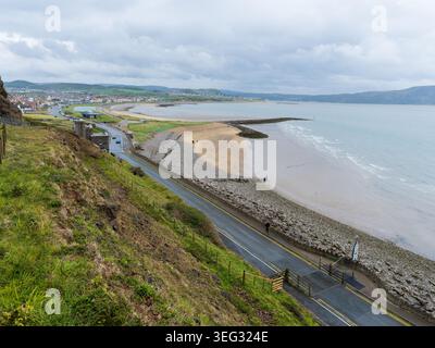 Great Orme coastal road winding along cliffs above West Shore beach, overlooking Llandudno, the Irish Sea and rocky peninsula under a cloudy, overcast Stock Photo