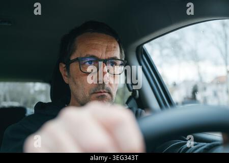 Focused middle-aged driver wearing glasses, hands on steering wheel and eyes fixed on road in serious concentration, selective focus Stock Photo