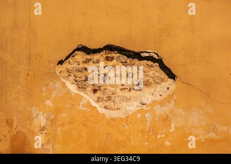 Weathered yellow plaster wall with peeling paint and exposed brick showing cracks, texture and decay; an architectural close-up conveying age, deterio Stock Photo