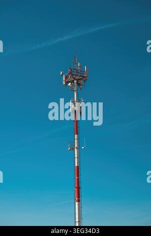 Red and white cell tower with antennas against clear blue sky for telecommunication and connectivity, vertical image. Stock Photo