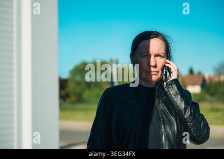 Serious middle-aged woman standing outdoors in a suburban area, wearing a black leather jacket while making a phone call on a mobile device under brig Stock Photo