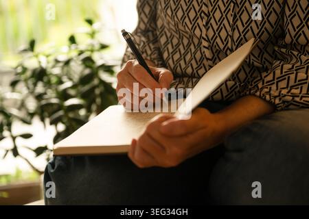 Middle-aged woman writing notes in notebook while sitting at home, reflective and productive evening scene, selective focus Stock Photo
