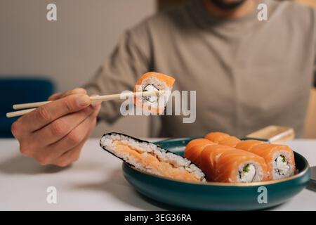 Close-up of unrecognizable person using chopsticks to pick philadelphia roll from dark green plate of salmon maki and cut-open rice roll, enjoying Stock Photo