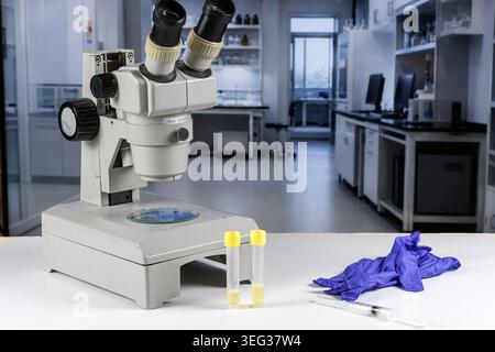 Microscope, sample tubes, syringe and protective gloves on a white bench in a clean lab, illustrating virus investigation and diagnostic testing. Stock Photo