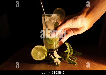 Hand is reaching for a tall glass holding a refreshing mojito cocktail with ice, mint leaves, and lime slices on a wooden surface, ready for a delicio Stock Photo