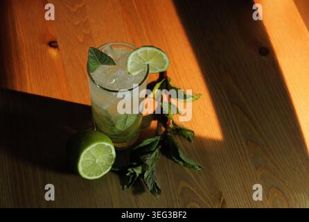 Mojito cocktail holding ice, lime slices, and fresh mint leaves in a tall glass, sitting on a wooden surface with a whole lime and mint sprig, illumin Stock Photo