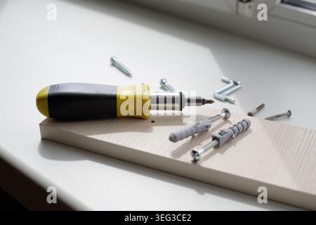 Screwdriver with a black and yellow plastic handle, dowels, and screws on a white window sill on a sunny day. DIY home improvement and repair concept Stock Photo