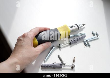 Hand picks up a screwdriver with a black and yellow rubber handle from a white windowsill. Dowels and screws are on the windowsill on a sunny day. Con Stock Photo