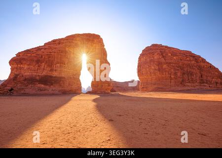 Elephant Rock (Jabal AlFil) towers in AlUla's sandstone desert as the setting sun creates a starburst through its natural arch, casting long golden Stock Photo