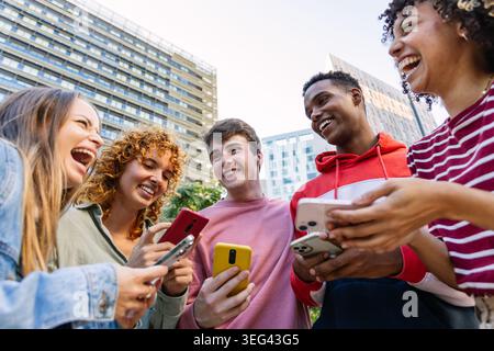 Diverse gen z friends laughing displaying smartphones outdoors Stock Photo