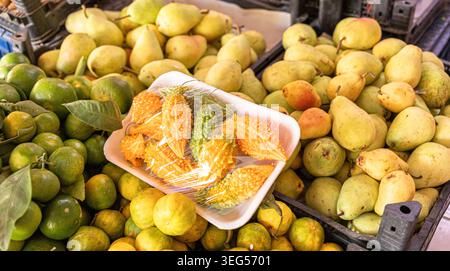 A variety of fresh fruits including green lemons, yellow lemons, and pears are arranged in a market setting, with a tray of wrapped bitter melons plac Stock Photo