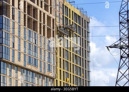 Workers are seen on a lift as they continue construction on the upper floors of buildings in a city under a blue sky with some clouds Stock Photo