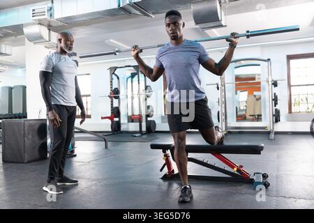 African American male trainer coaching client doing split squat with barbell on bench at gym Stock Photo
