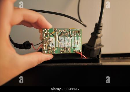 Close up of a damaged electronic circuit board held in hand near power cables indoors. Burnt electrical component and technology repair concept. Stock Photo