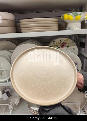 A hand holds a speckled beige dinner plate in front of a shelf filled with various dishes and decorative items Stock Photo