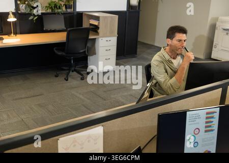 Studying analyst at cubicle in olive shirt holding pen, viewing monitor showing graphs, copy space. Desk, workstation, openplan, carpet, plants, focus Stock Photo
