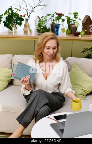 Relaxing in her serene living space, a mature woman engages with her journal while savoring coffee. Stock Photo