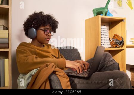 Black teenager wearing headphones and using laptop while studying from home, showing online learning and digital focus. Useful for education technology, remote school, youth learning campaigns Stock Photo